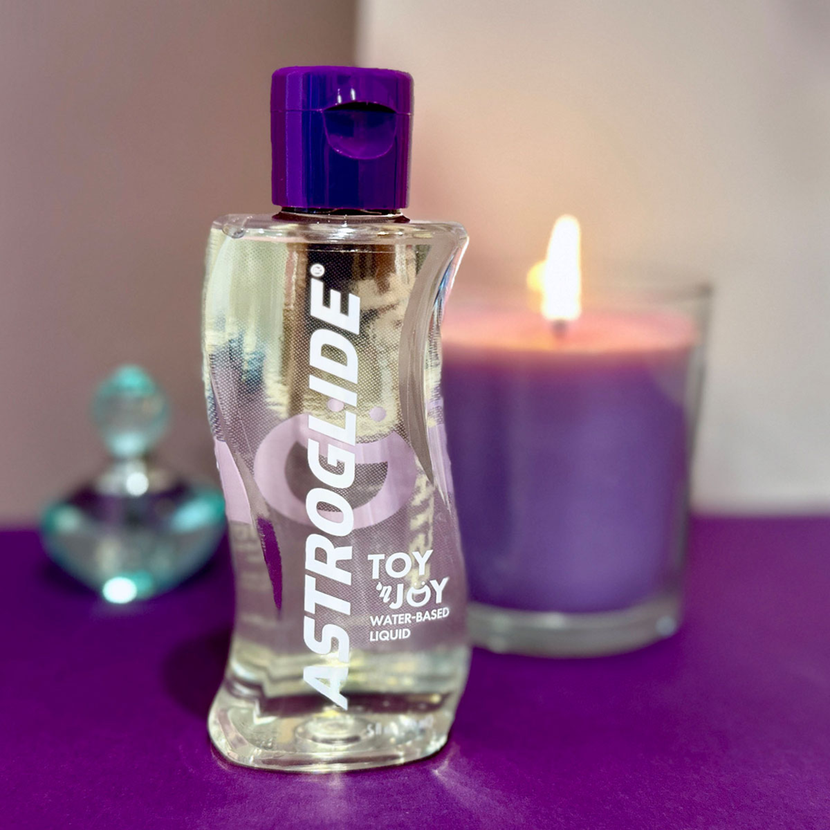 A bottle of Astroglide Toy 'N Joy Lubricant with a purple cap sitting on a purple surface, next to a lit purple candle.