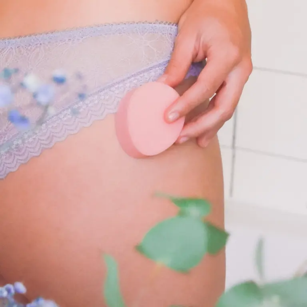 An image showing a person's hand holding a pink sponge tampon against their upper thigh. The person is wearing light purple lace underwear. The background is soft-focused with hints of green leaves and white tiles, suggesting a bathroom or personal care setting.
