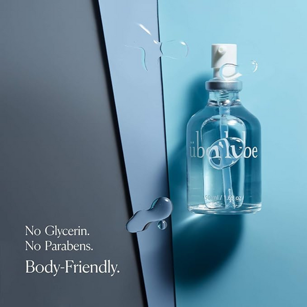 A clear glass bottle of Überlube lubricant, with a white spray top, stands upright on a blue surface, while a dark gray panel diagonally cuts through the left side of the frame. Droplets of clear liquid are visible on both surfaces, emphasizing the product's silky texture, while the text "No Glycerin. No Parabens. Body-Friendly." highlights key benefits of this trusted WorldCondoms offering.