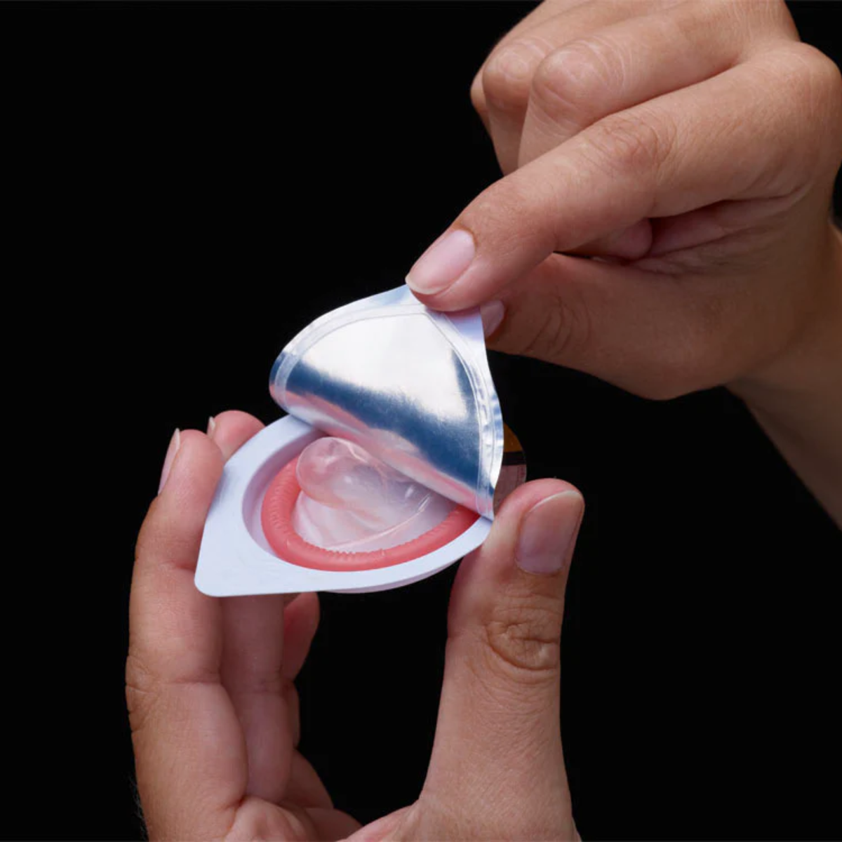 Hands opening a single foil packet to reveal a rolled, lubricated condom. 