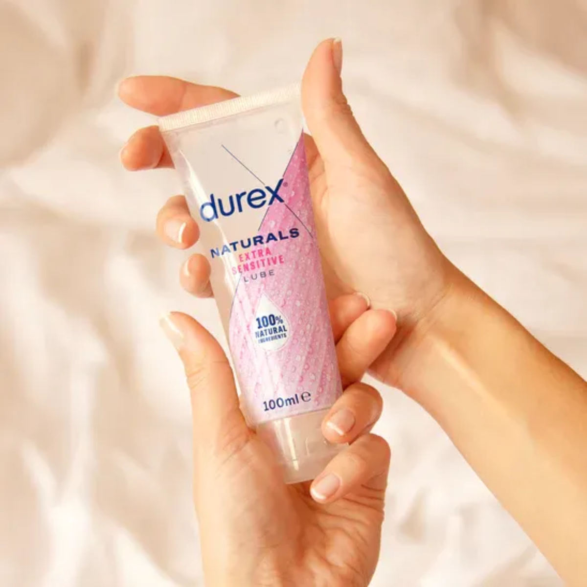 A pair of hands holding a tube of Durex Naturals Extra Sensitive Lube. The shot is taken against a warm, neutral fabric background to showcase the product's size and portable design.