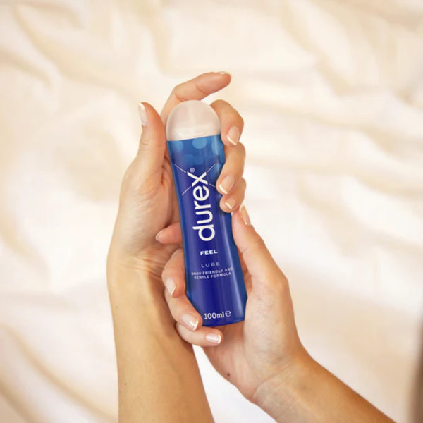 A pair of hands holding a dark blue Durex Feel Lube bottle against a background of white sheets. The bottle is labeled "FEEL LUBE BODY-FRIENDLY AND GENTLE FORMULA."