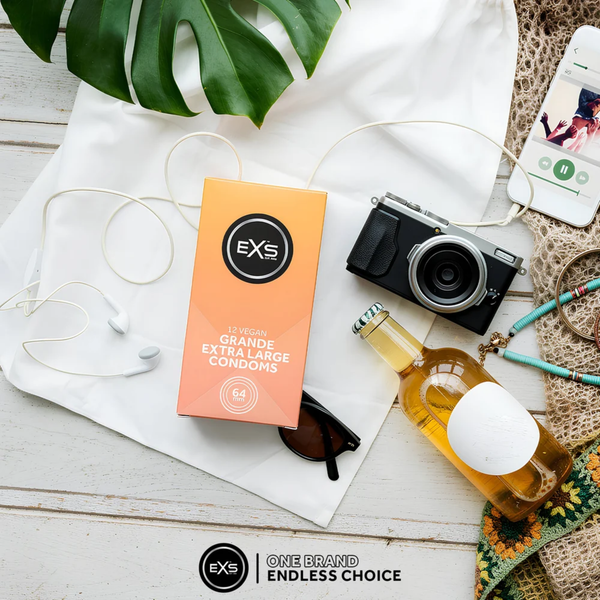 Top-down flat lay view of a peach-colored box of EXS 12 Vegan Grande Extra Large Condoms displayed among travel accessories, including a vintage camera, white earphones, sunglasses, a beverage bottle, and a smartphone on a white wooden surface.