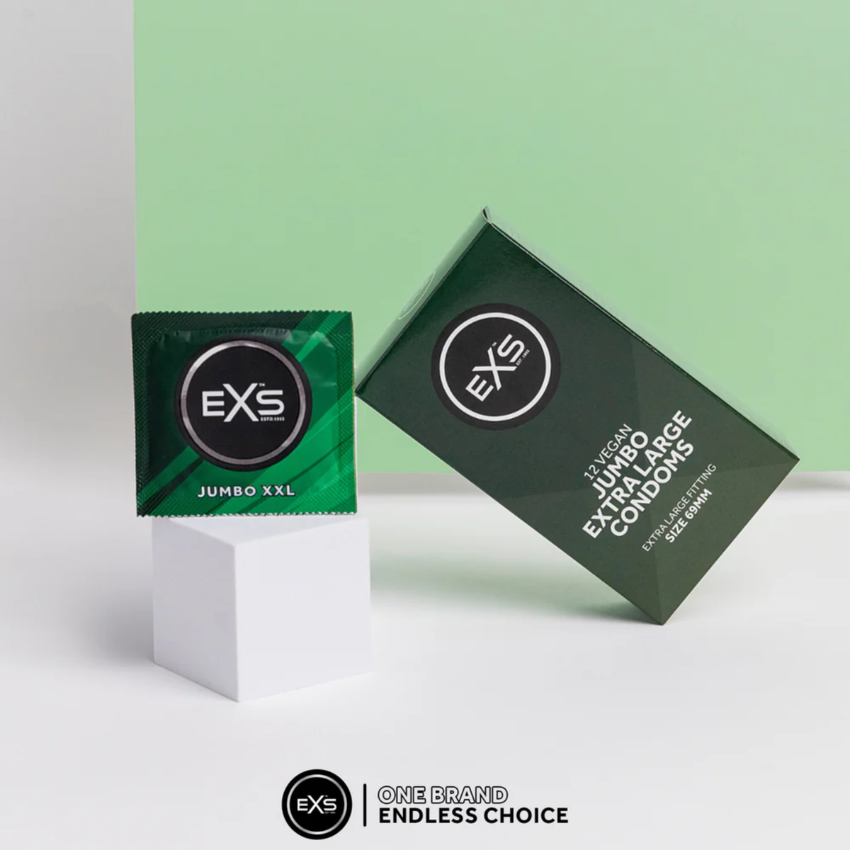 Product display showing a single EXS Jumbo XXL foil wrapper standing upright on a white cube, next to the dark green EXS 12 Vegan Jumbo Extra Large Condoms box resting at an angle against a split light green and white background.