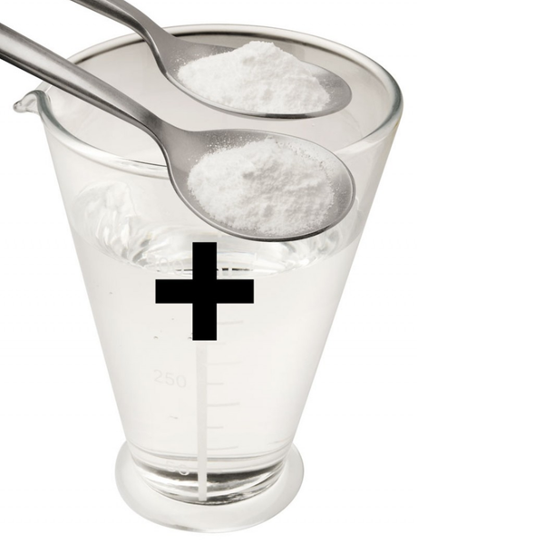 An instructional graphic demonstrating how to mix the product. Two silver spoons filled with fine white powder are held over a clear glass measuring beaker filled with water. A large black plus sign (+) is overlaid on the beaker, indicating that the powder should be added to the liquid to create the lubricant.
