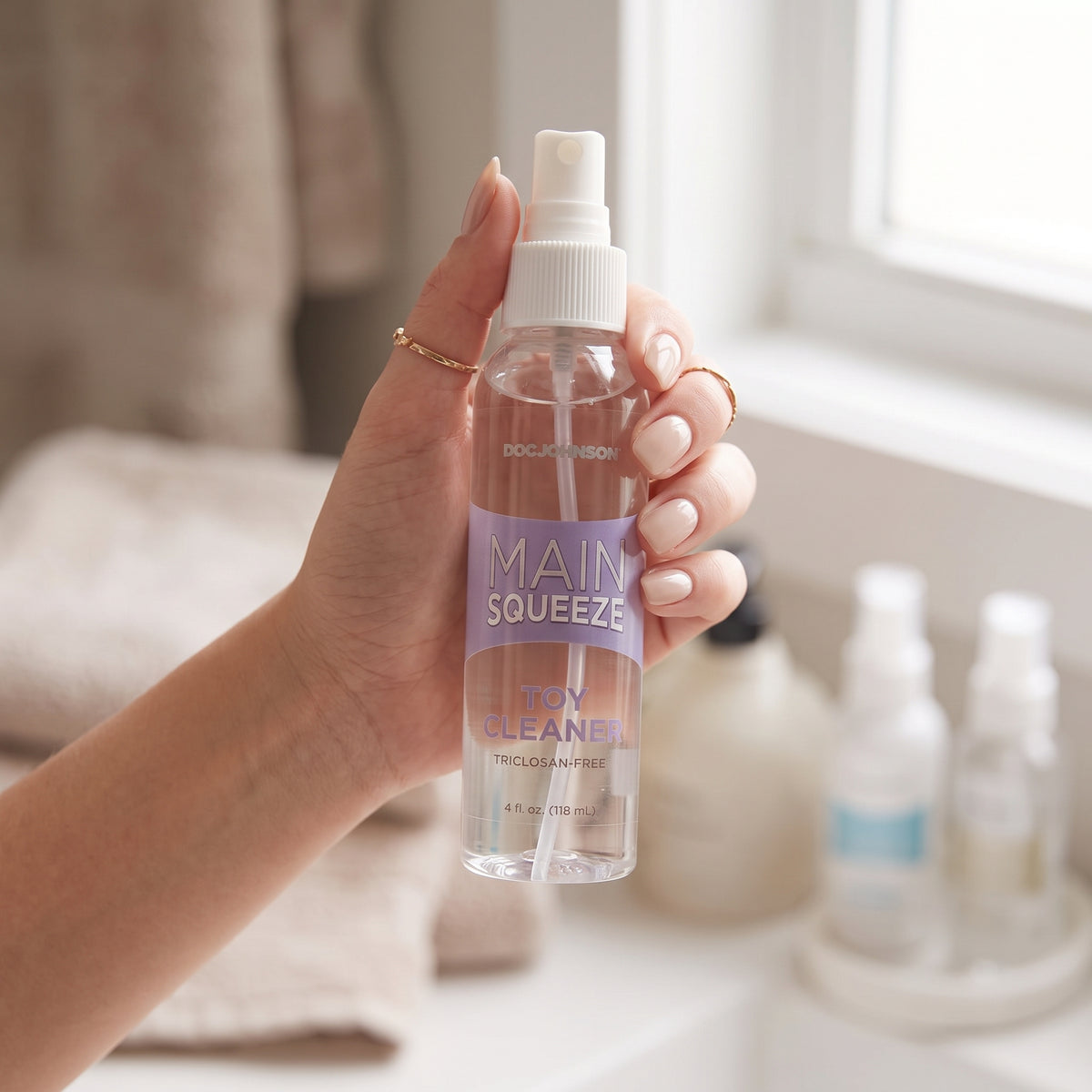 Lifestyle shot showing a person's hand holding the 4 fl oz (118 ml) clear spray bottle of Doc Johnson Main Squeeze Toy Cleaner in a brightly lit bathroom setting.