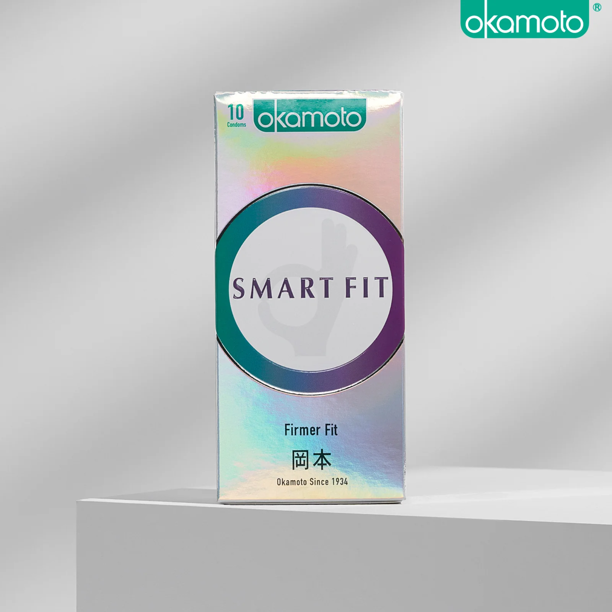 A product photo of a rectangular, holographic Okamoto 'SMART FIT' condom box containing 10 condoms. The box features a purple and green ring design and is placed on a white surface with a gray background. The text "Firmer Fit" is visible.