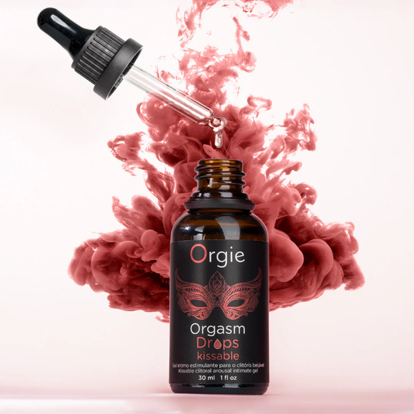 A 30ml amber bottle of Orgie Orgasm Drops centered against a light pink background. A glass dropper is angled above the open bottle with a single clear drop falling into it. Directly behind the bottle, a vibrant, billowy cloud of reddish-pink ink or smoke explodes outward, creating a dynamic and sensual visual effect. The label identifies the product as a "Kissable clitoral arousal intimate gel".