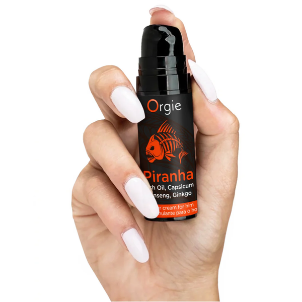 A close-up of a person with long white manicured nails holding the compact 15ml bottle of Orgie Piranha Power Cream. The cap is removed to reveal a black pump-action dispenser, showing the small, portable size of the product.
