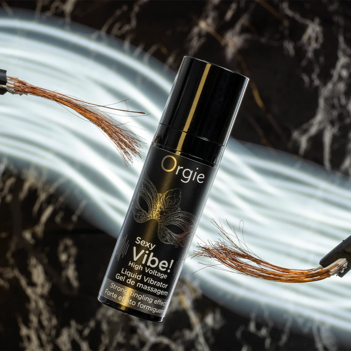 A small black pump bottle of Orgie Sexy Vibe! High Voltage Liquid Vibrator Gel with a gold mask design, placed on a dark marble surface surrounded by glowing white light cables and frayed copper wires, symbolizing the strong tingling and vibrating effect.