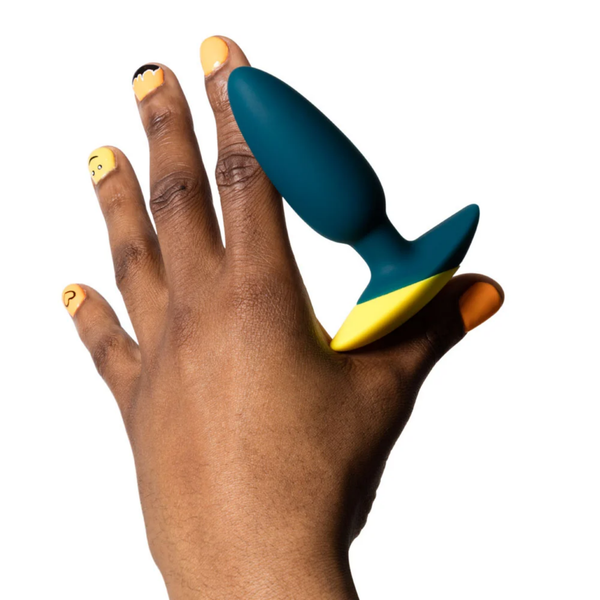 Scale perspective photo showing a hand holding the dark teal and yellow ROMP Bass vibrating anal plug between the fingers.