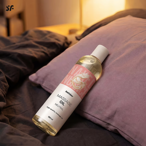 Lifestyle shot of the Satisfyer Naked Nuance Neutral Massage Oil bottle resting against a purple pillow on a dimly lit bed.