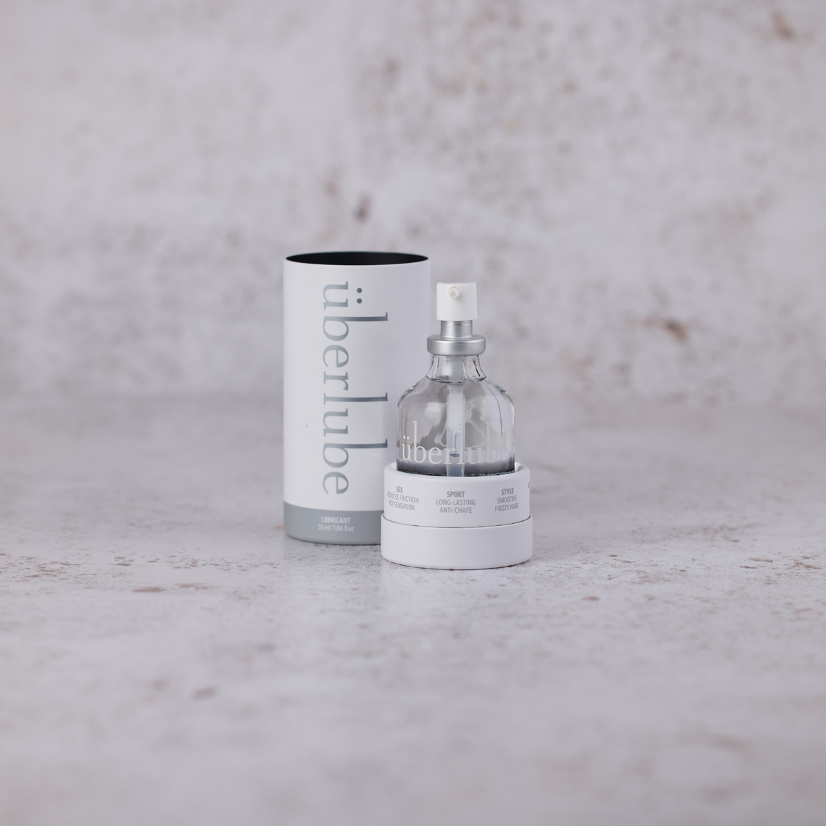 A small, clear glass bottle of Überlube lubricant, with a silver pump and a white base, stands uncapped on a light gray, textured surface. To its left, the white cylindrical outer packaging tube, bearing the "überlube" logo, stands upright. The background is mottled light brown and white color providing a neutral backdrop to the product. From worldcondoms assortment.