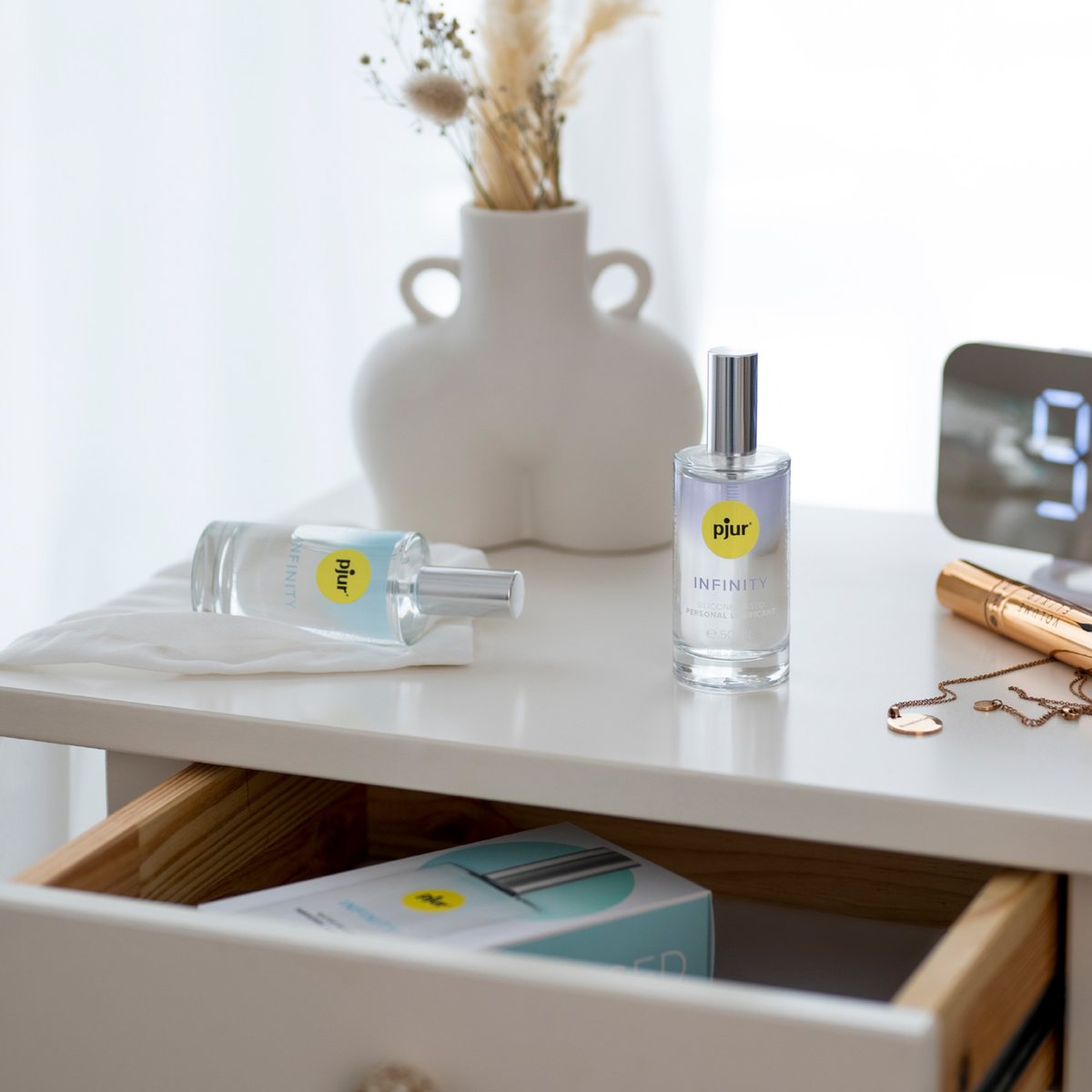 A photo of two bottles of pjur Infinity Water-Based Personal Lubricant sitting on a white nightstand. One bottle is standing upright, and the other is lying on its side on a white cloth.