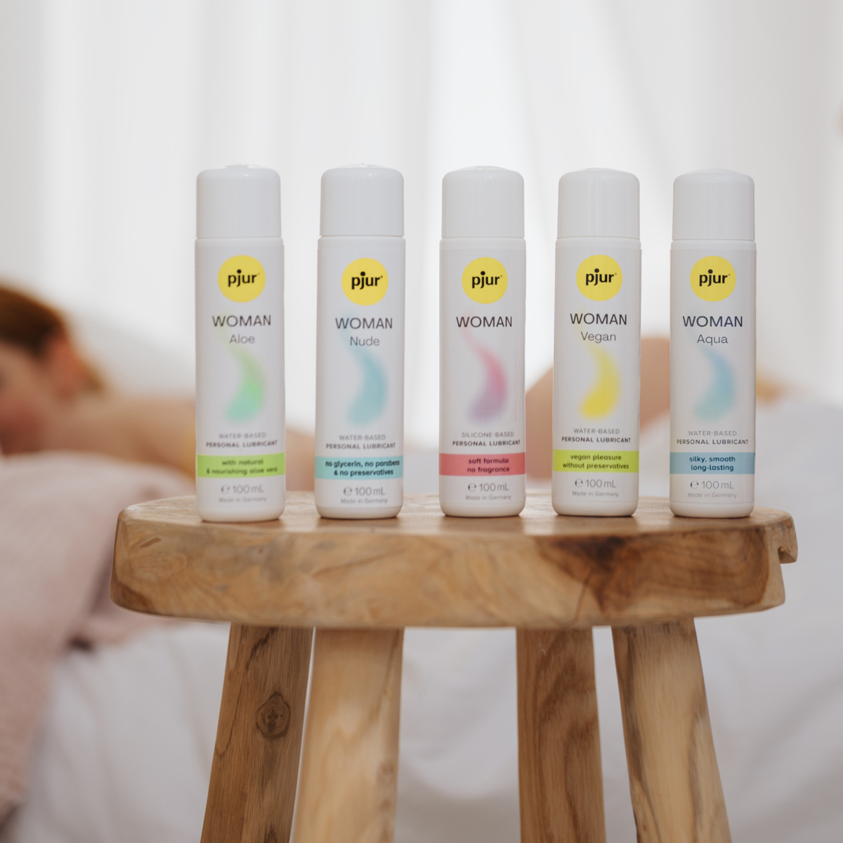 Five bottles of pjur Woman personal lubricants (Aloe, Nude, Nude, Vegan, Aqua) sitting on a small wooden stool, with a person relaxing in the blurred background.