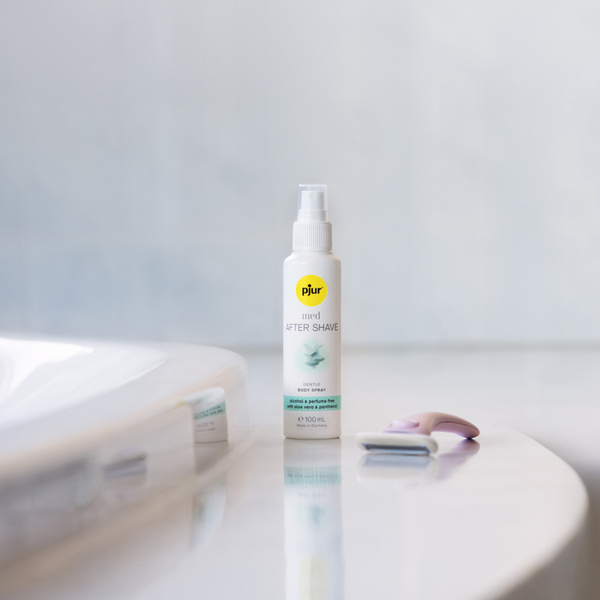 A white bottle of pjur med AFTER SHAVE spray stands on a white surface next to a pink and grey razor, with a blurred white object in the foreground and a light grey background.