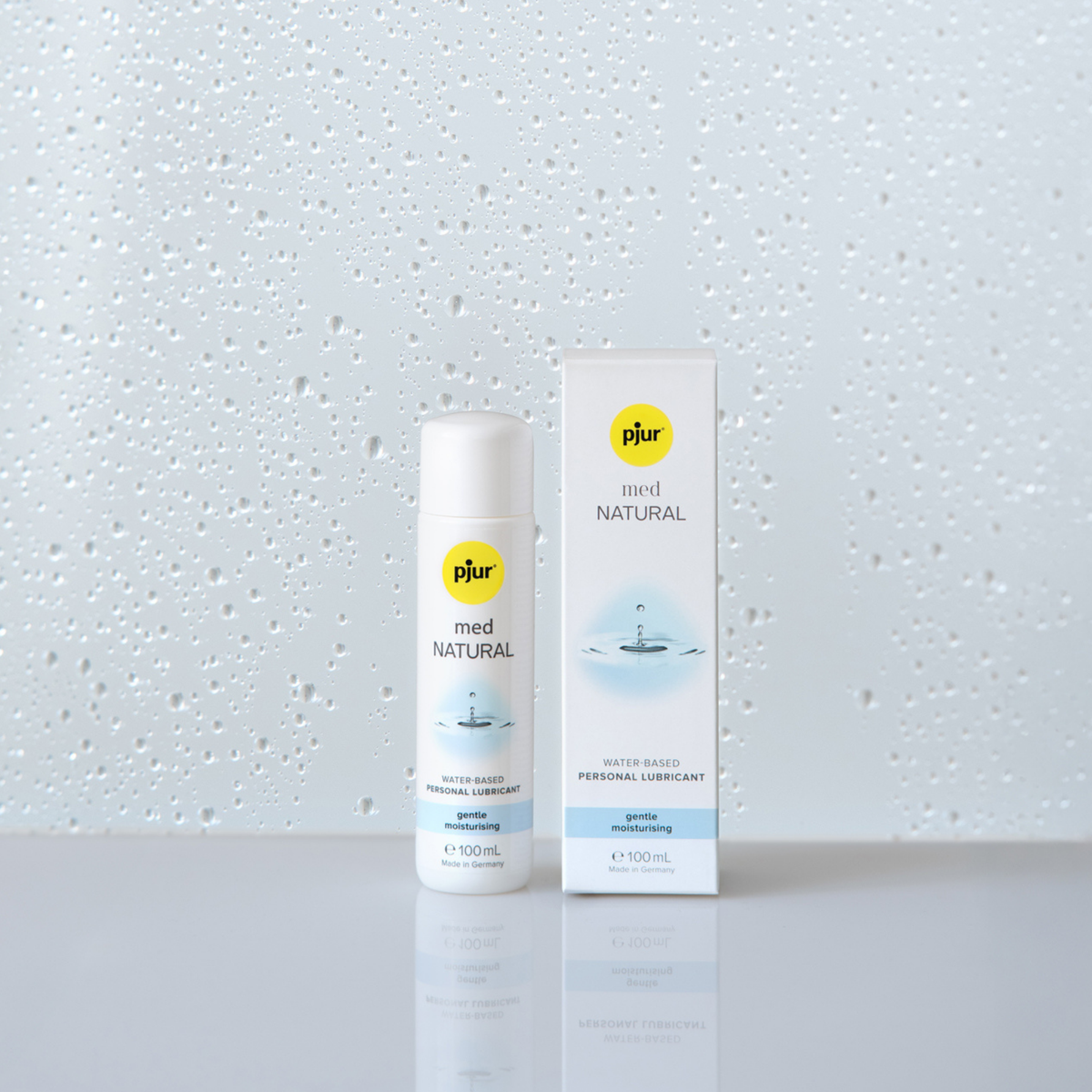 A white bottle and box of pjur med NATURAL water-based personal lubricant stand on a white surface against a background with water droplet patterns.