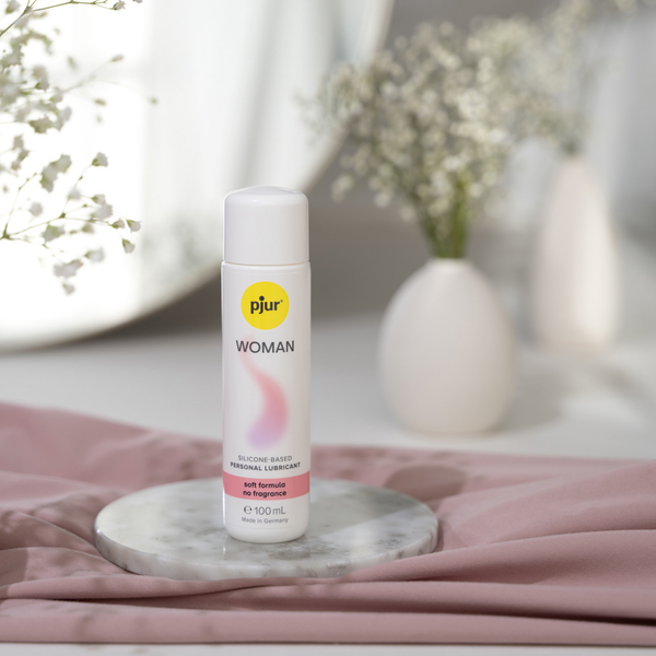 A bottle of Pjur Woman silicone-based personal lubricant on a marble coaster with a blurred background of flowers and fabric.