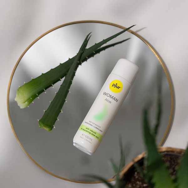 A white bottle of Pjur Woman Aloe water-based personal lubricant lies on a round gold-rimmed mirror next to two cut aloe vera leaves. 