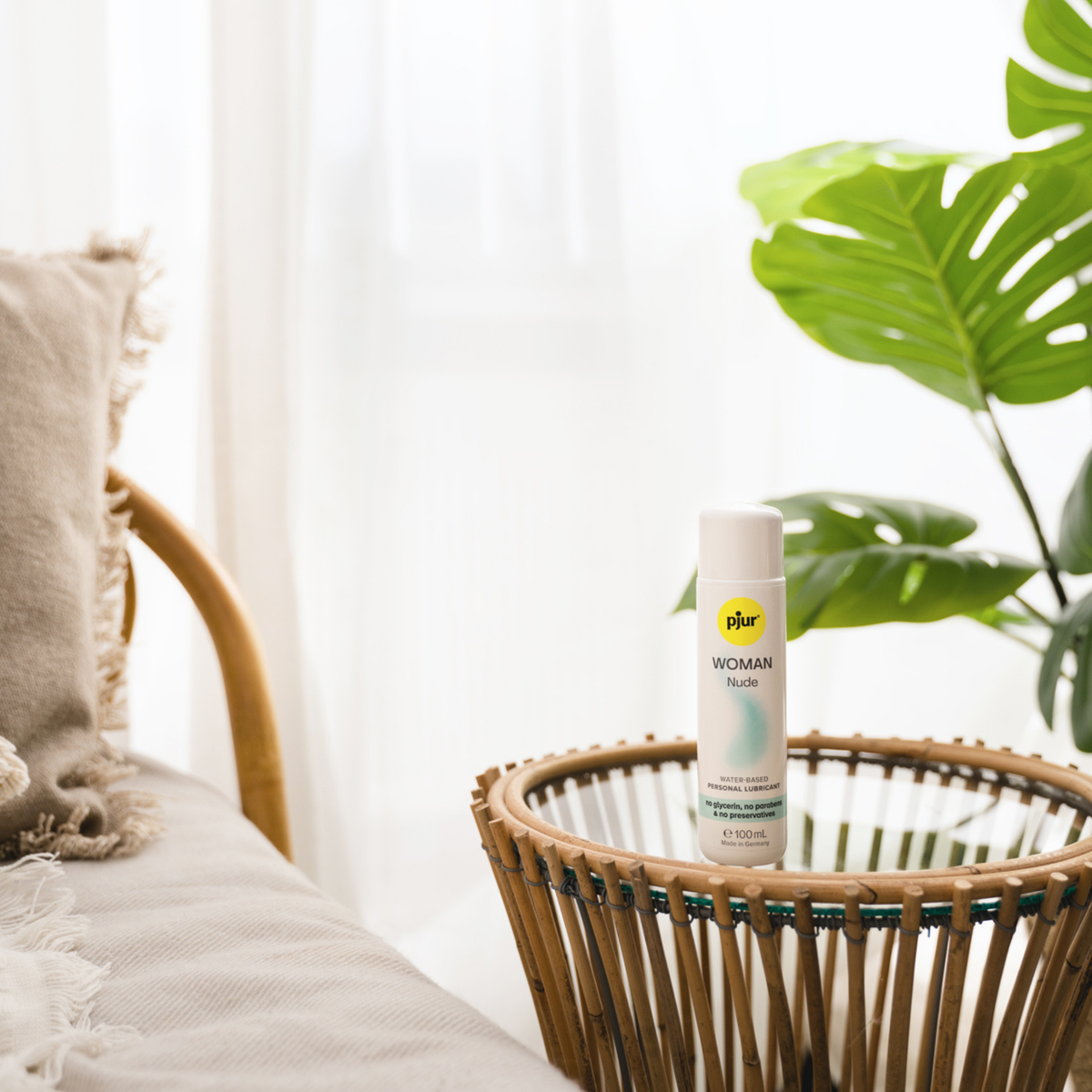 A white bottle of pjur WOMAN Nude water-based personal lubricant sits on a round wicker table next to a chair and a potted plant.