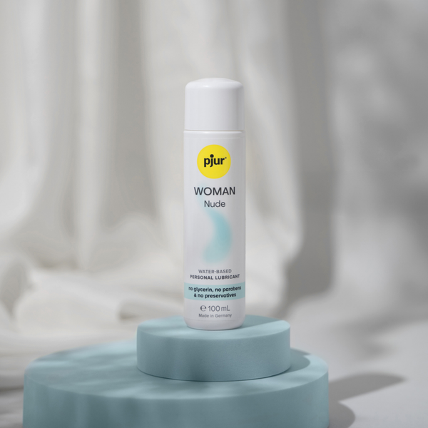 A white bottle of pjur WOMAN Nude water-based personal lubricant standing on a light blue geometric platform against a soft, blurred background.