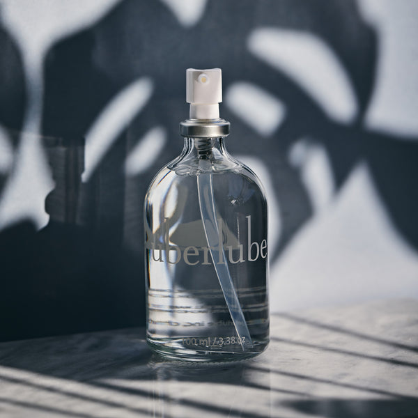 
A clear glass bottle of Überlube lubricant, with its spray top, stands prominently against a textured dark background, casting a distinct shadow of a plant. This sleek product, available through WorldCondoms, is designed for smooth and lasting personal comfort