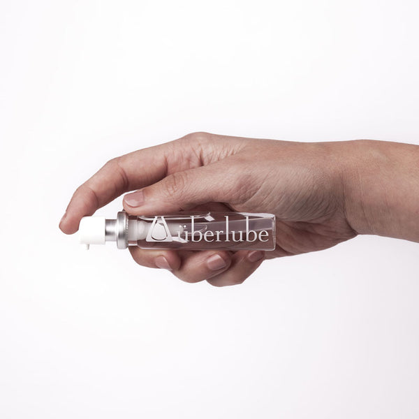 A hand holds a clear, horizontal Überlube lubricant spray bottle, prominently displaying the "überlube" logo. This compact WorldCondoms product offers discreet and convenient personal lubrication.